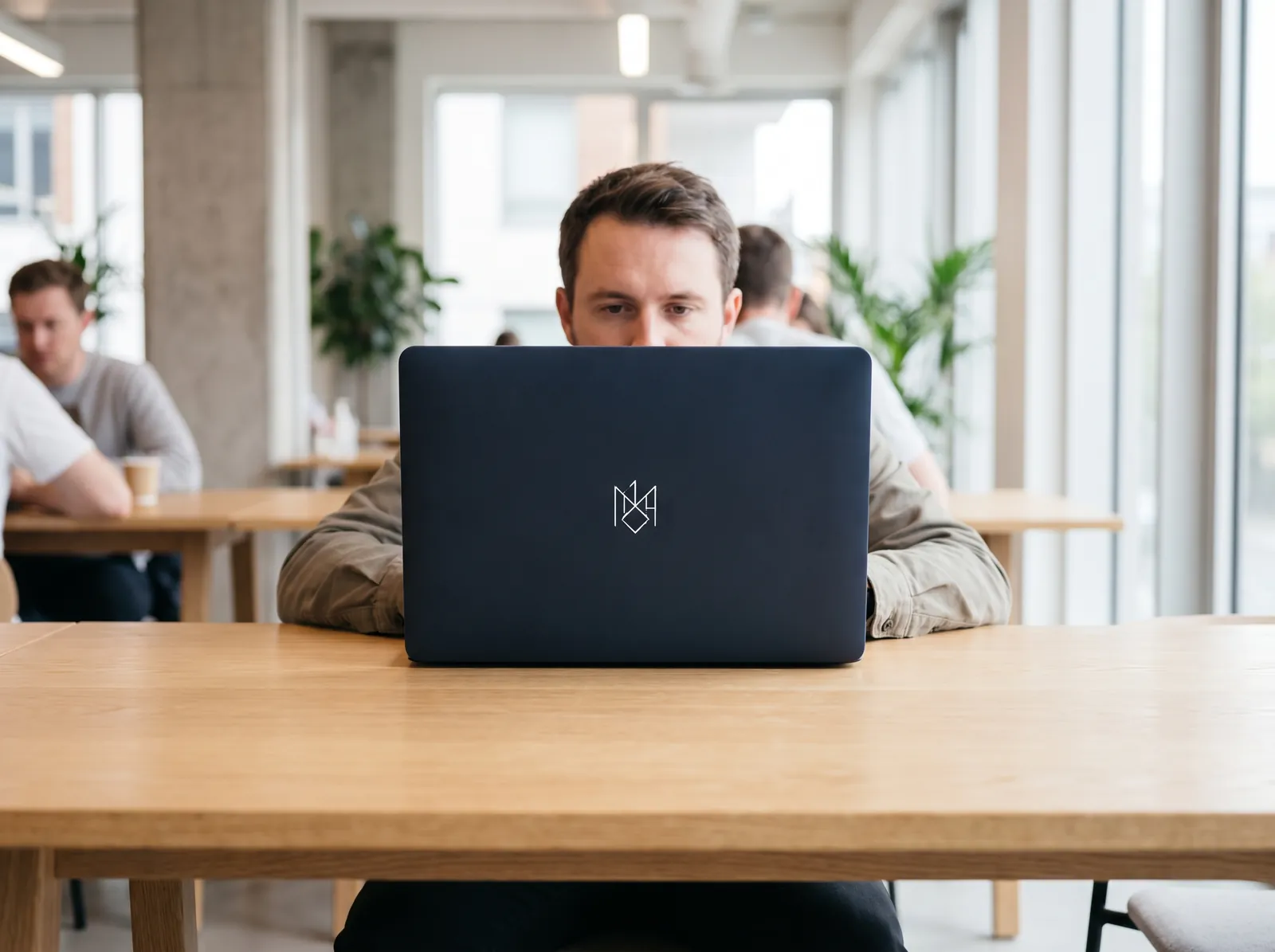 A founder working at a coworking table in a coworking space, the back of their laptop showing a navy custom cover with a white monogram
